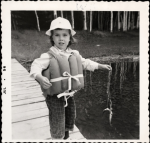 Child in a life jacket holding fish on a dock in July 1956